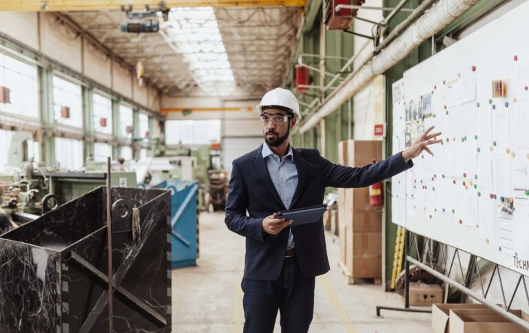 A man in a hardhat pointing at a white board in a manufacturing plant.